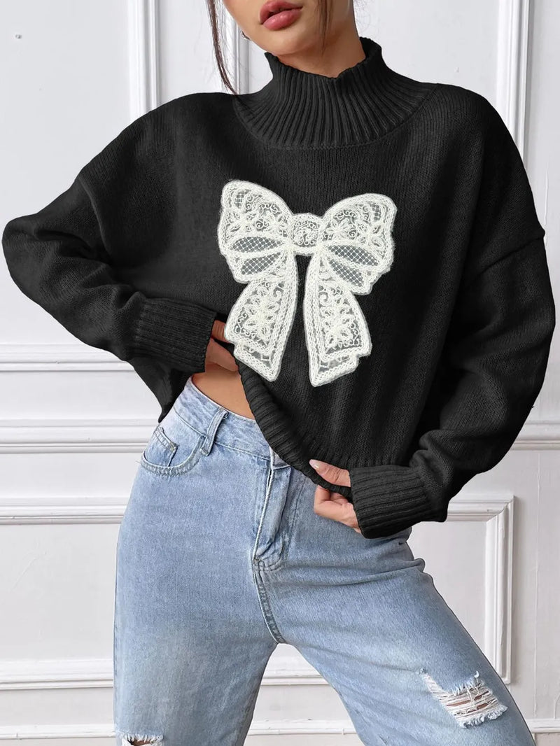 Person wearing a black sweater with a large white bow design, standing against a white wall.