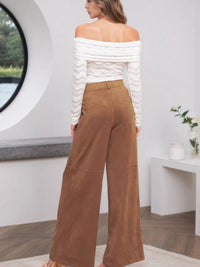 Woman wearing a white off-shoulder top and brown wide-leg pants in a modern room.