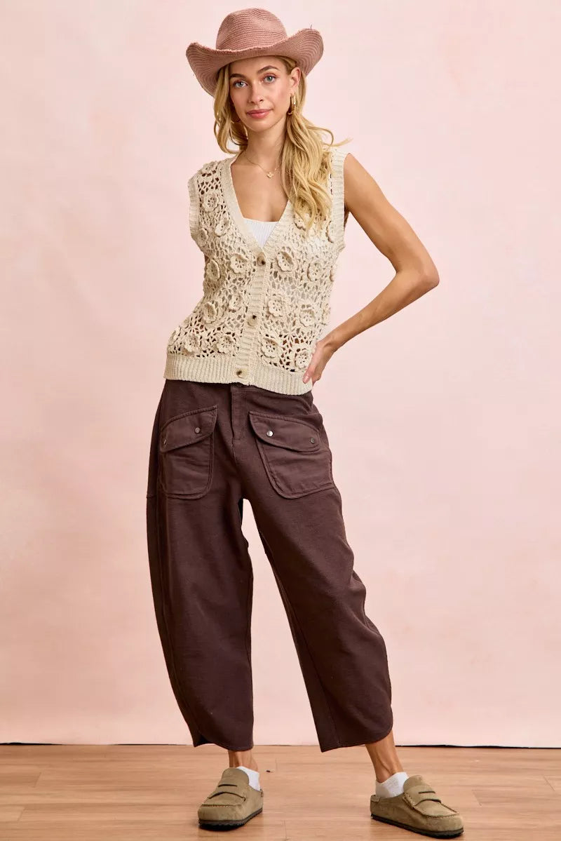 Woman wearing a beige sleeveless top, brown wide-leg pants, and a pink cowboy hat against a pink background.