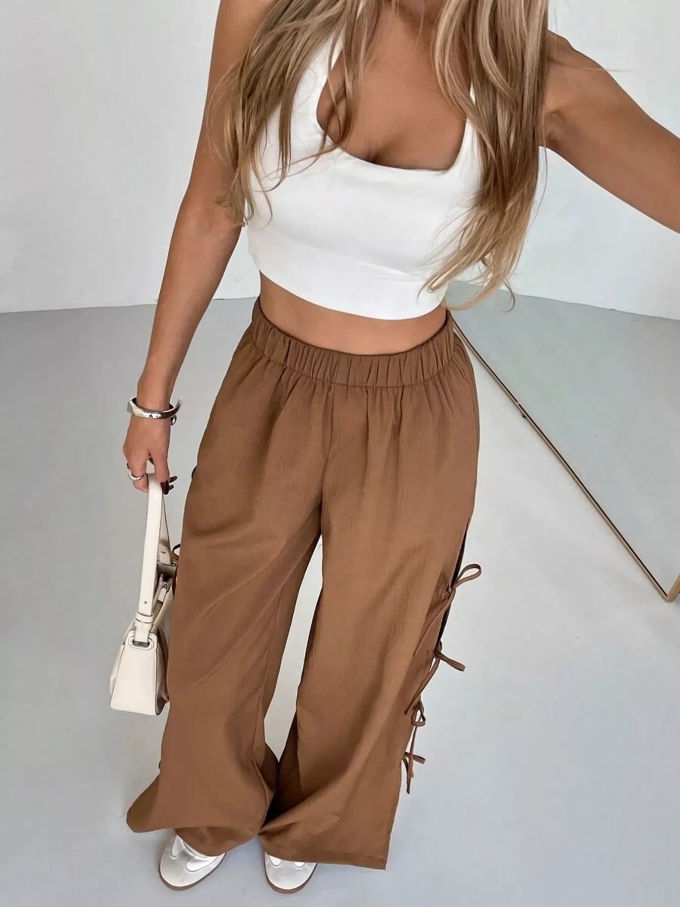 Woman wearing a white crop top and brown wide-leg pants, holding a beige handbag.