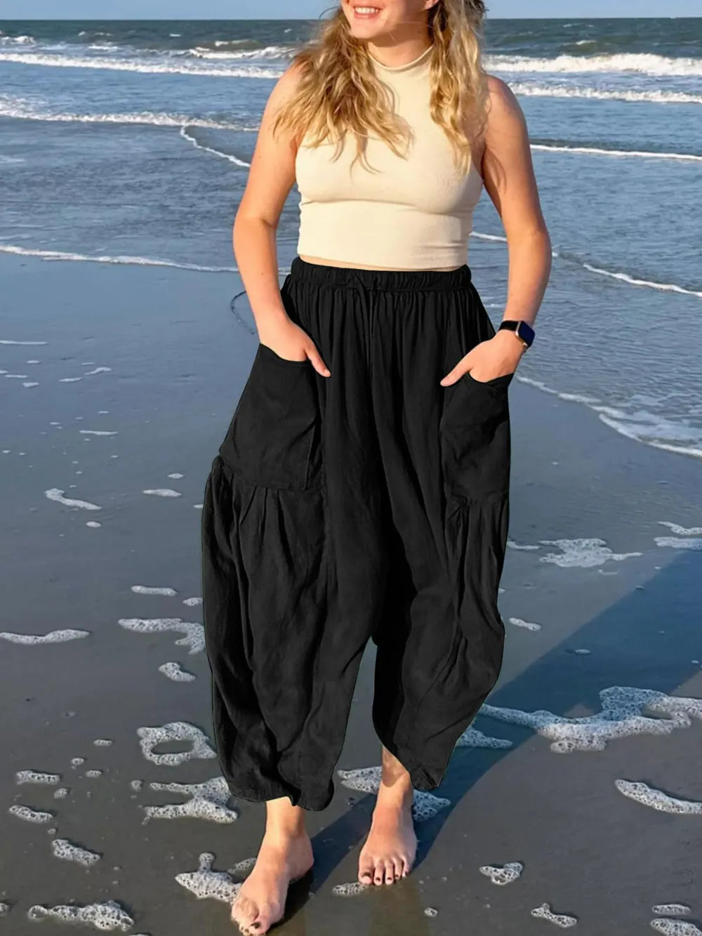 Woman standing on a beach wearing a beige top and black harem pants.