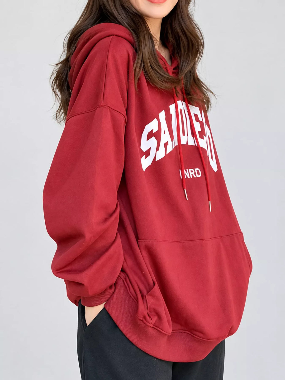 A woman wearing an oversized red hoodie with white 'SAIDEN' and 'I N R D' text on the front, paired with black pants, against a light gray studio background.