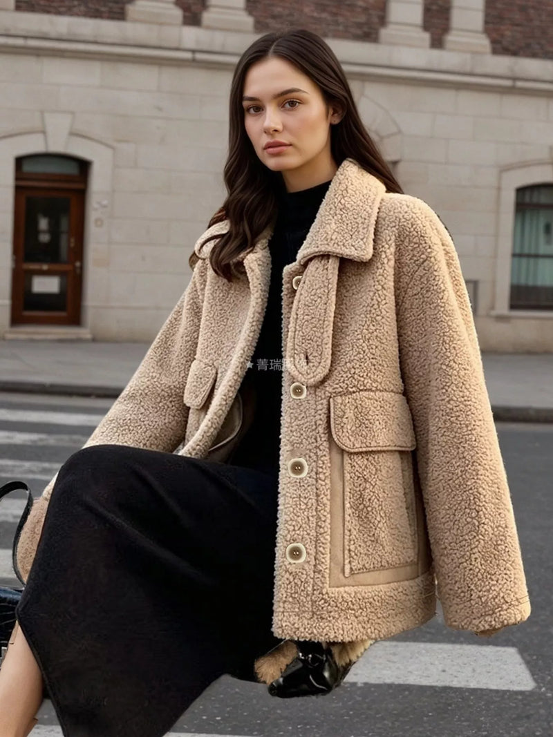 A woman wearing a beige sherpa fleece jacket with large front pockets and a wide collar, styled over a black top and black skirt, sitting on a city street crosswalk.