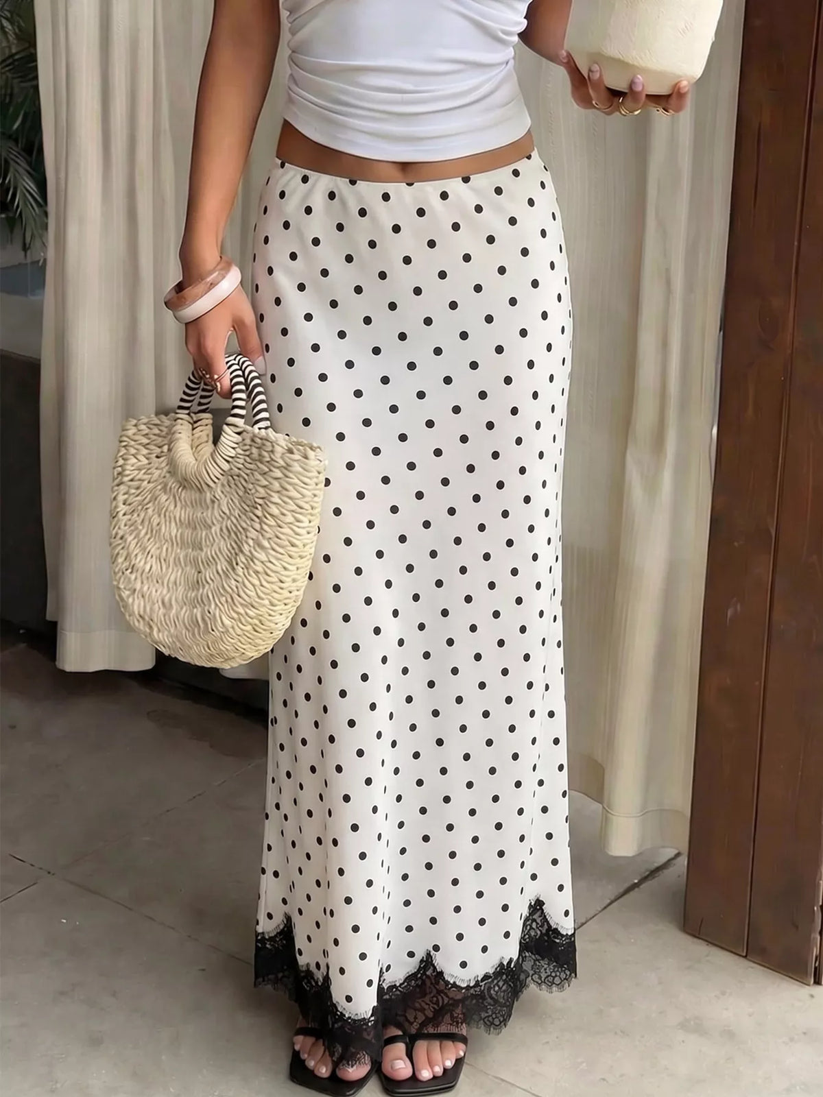 A woman wearing a long white maxi skirt with black polka dots and a black lace hem, styled with a white crop top, black sandals, a woven straw bag, and holding a coconut in a home setting.