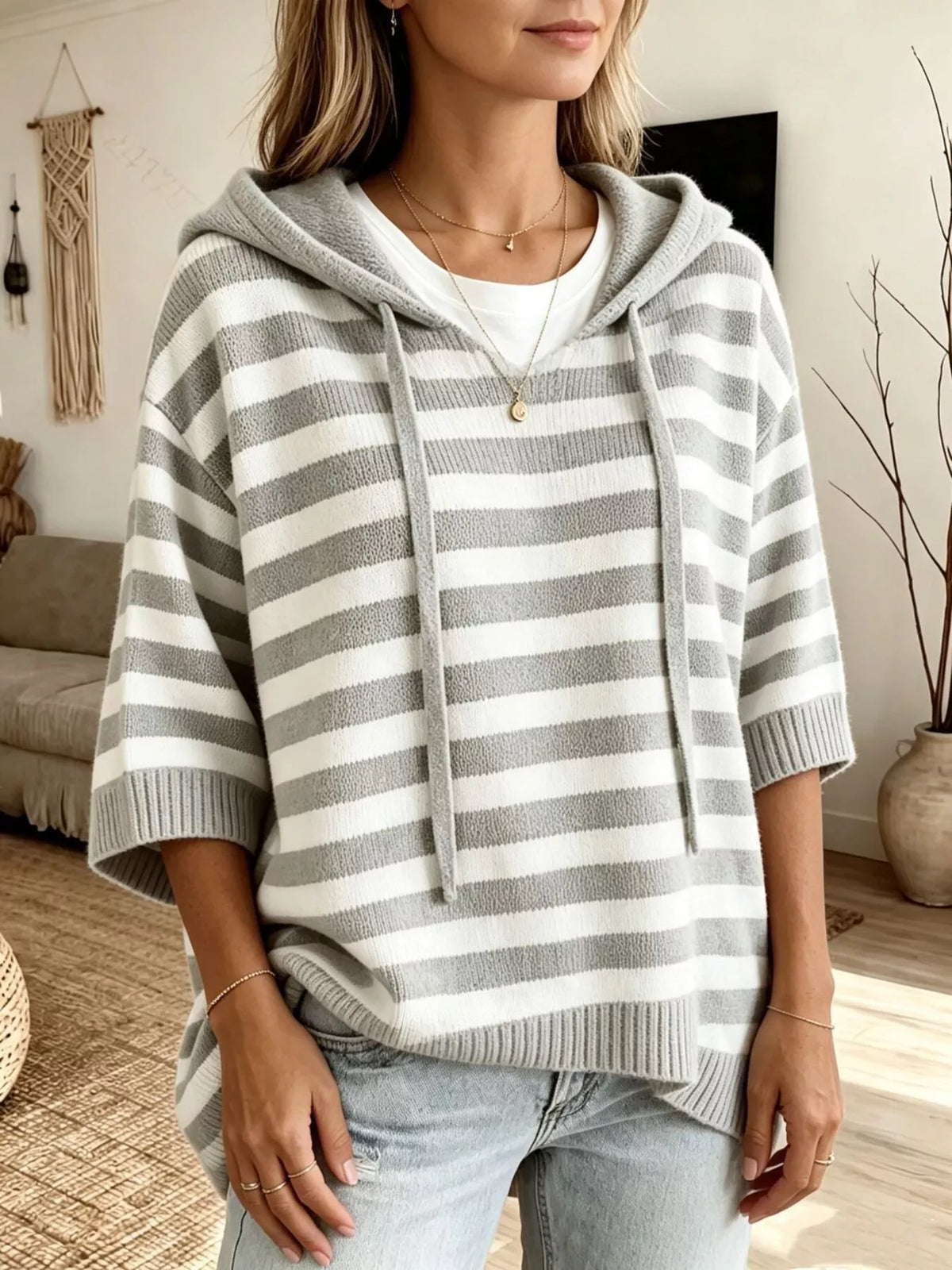 A woman wearing a gray and white horizontally striped knit hoodie with drawstrings, layered over a white t-shirt and paired with light-wash jeans in a cozy indoor setting.
