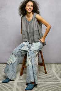 Woman sitting on a stool wearing a sleeveless top and patchwork jeans against a gray background