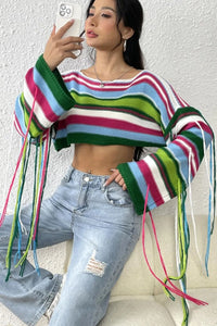 Woman wearing a colorful striped sweater with tassels and ripped jeans, sitting on a white chair.