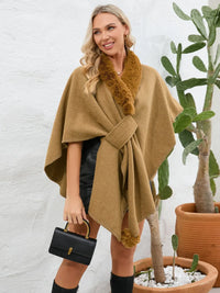 Woman wearing a brown poncho with fur trim, holding a black handbag, standing next to potted plants.