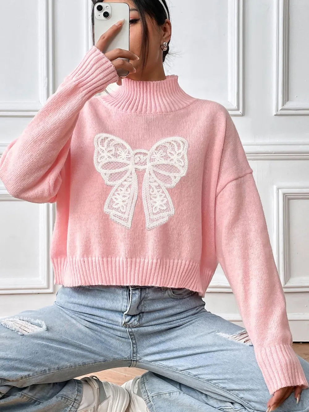 Person wearing a pink sweater with a white bow design, sitting against a white paneled wall.