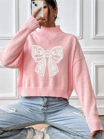 Person wearing a pink sweater with a white bow design, sitting against a white paneled wall.