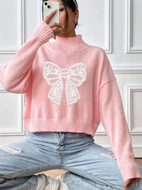 Person wearing a pink sweater with a white bow design, sitting against a white paneled wall.