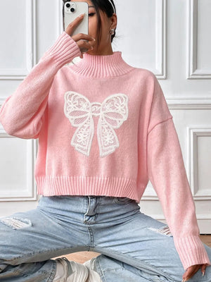 Person wearing a pink sweater with a white bow design, sitting against a white paneled wall.
