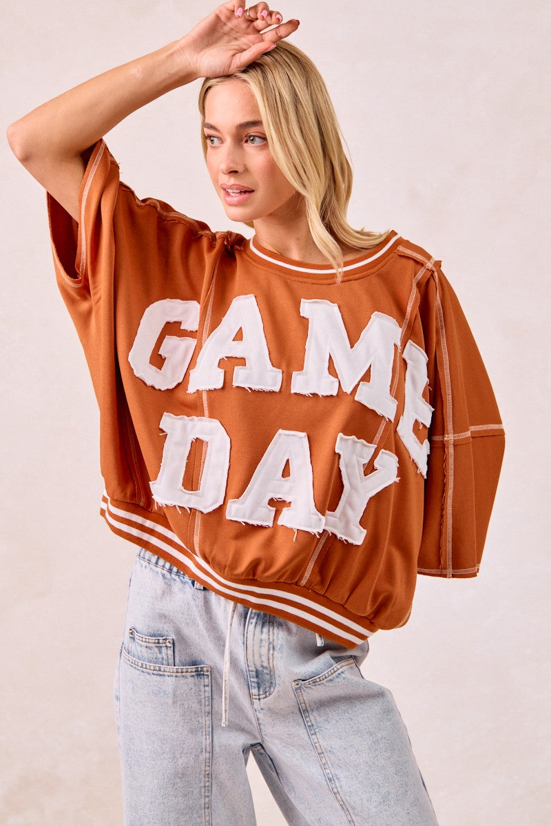 Woman wearing a brown 'GAME DAY' shirt with light blue jeans on a beige background