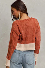 Woman wearing a rust-colored sweater over a beige top with blue jeans on a gray background