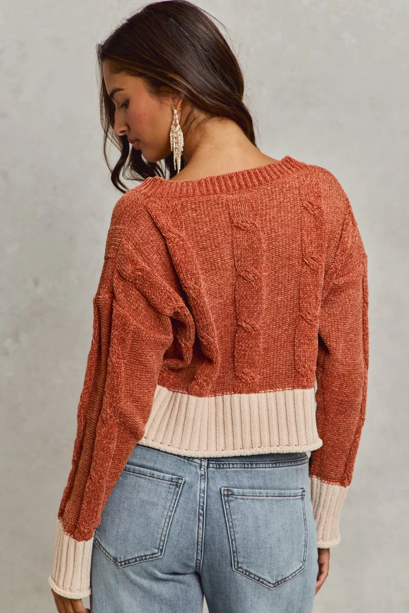 Woman wearing a rust-colored sweater over a beige top with blue jeans on a gray background