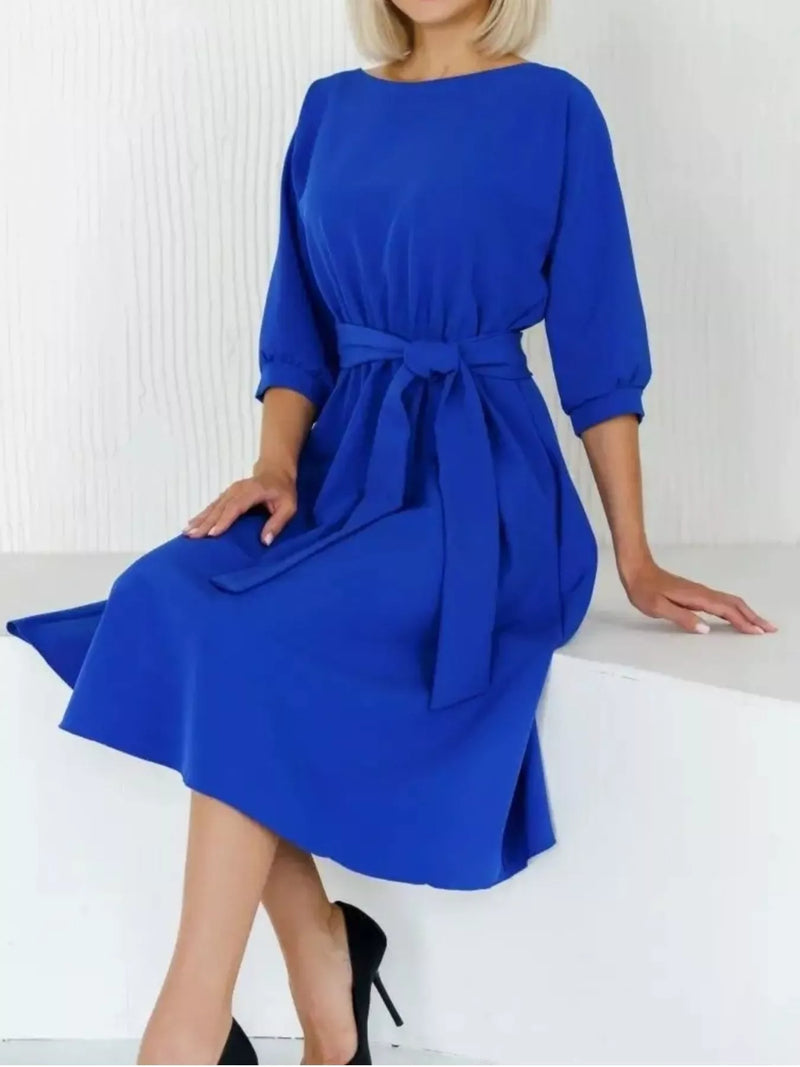 A woman wearing a bright royal blue knee-length dress with three-quarter sleeves and a matching waist tie, seated on a white ledge in a clean indoor setting.