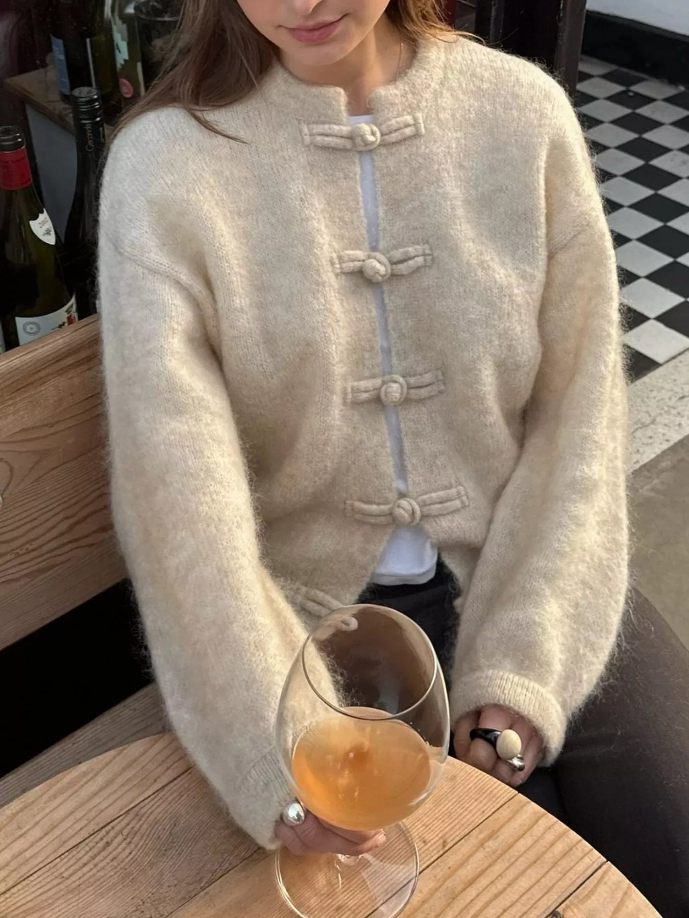 Person wearing a beige sweater holding a glass of orange liquid in a casual setting.