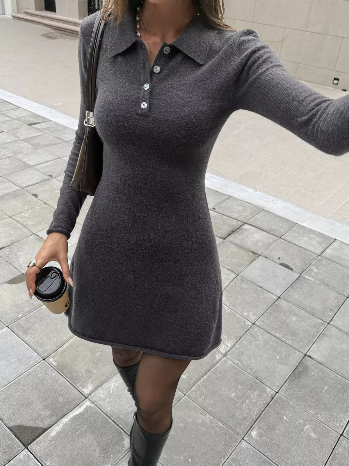 A woman wearing a fitted long-sleeve gray knit polo dress with a buttoned collar, standing outdoors on a paved walkway and holding a takeaway coffee cup.