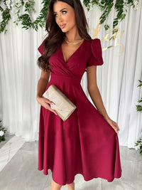 Woman in a burgundy dress holding a clutch against a white curtain background