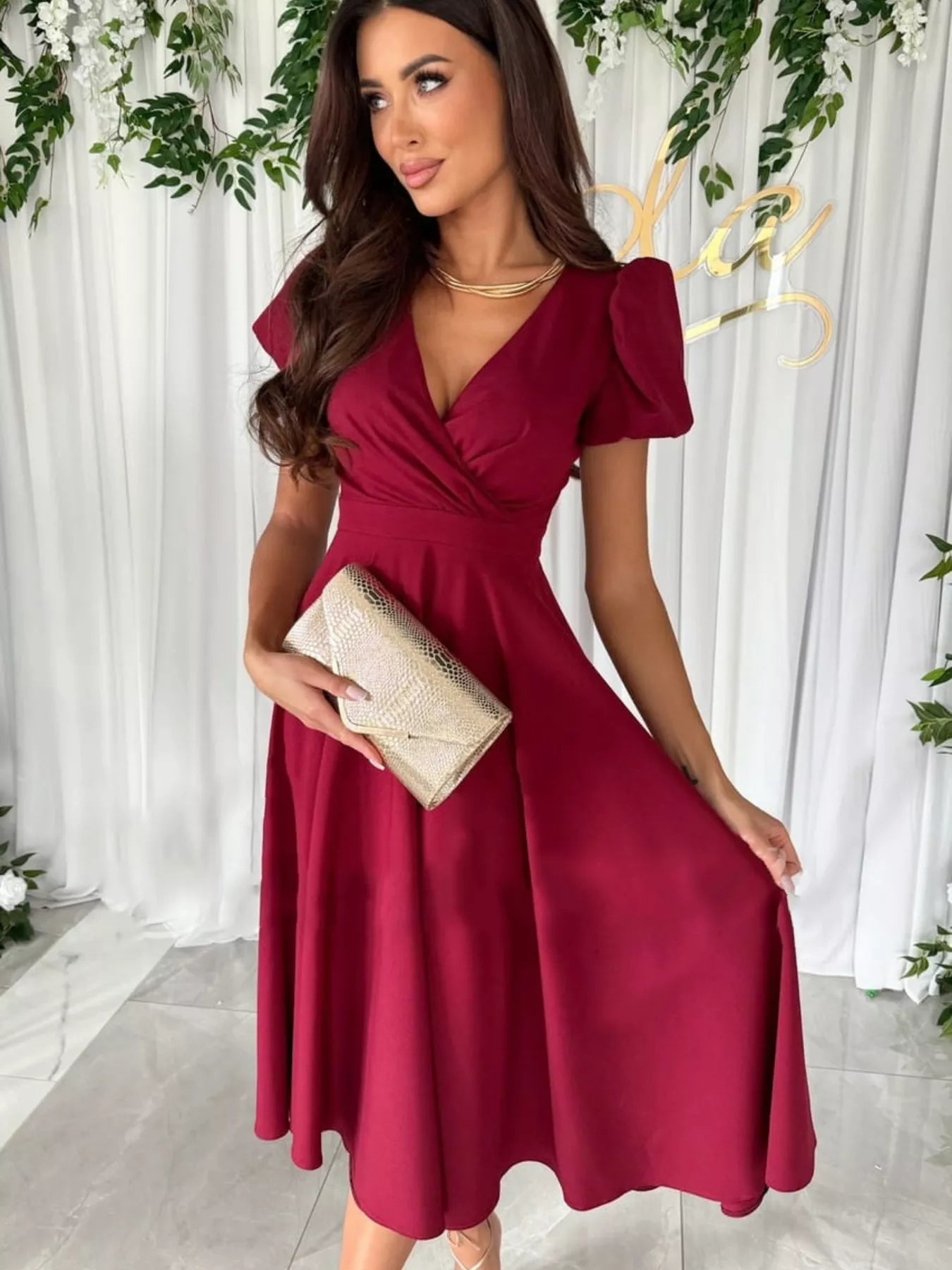 Woman in a burgundy dress holding a clutch against a white curtain background