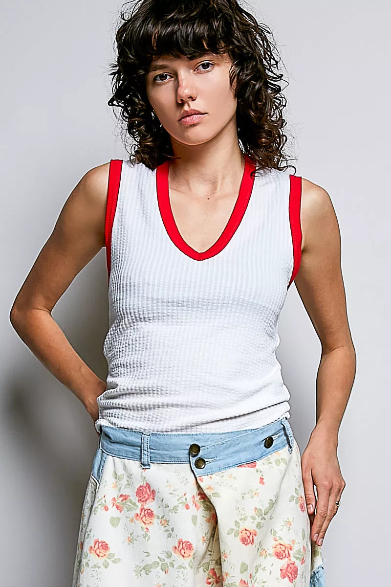 Woman wearing a white tank top with red trim and floral pants against a plain background
