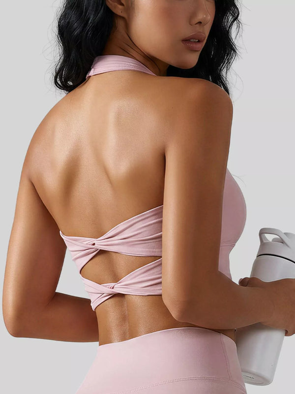 A woman shown from the back wearing a light pink athletic halter crop top with a knotted crossover back detail, paired with matching pink bottoms, against a plain light gray studio background.