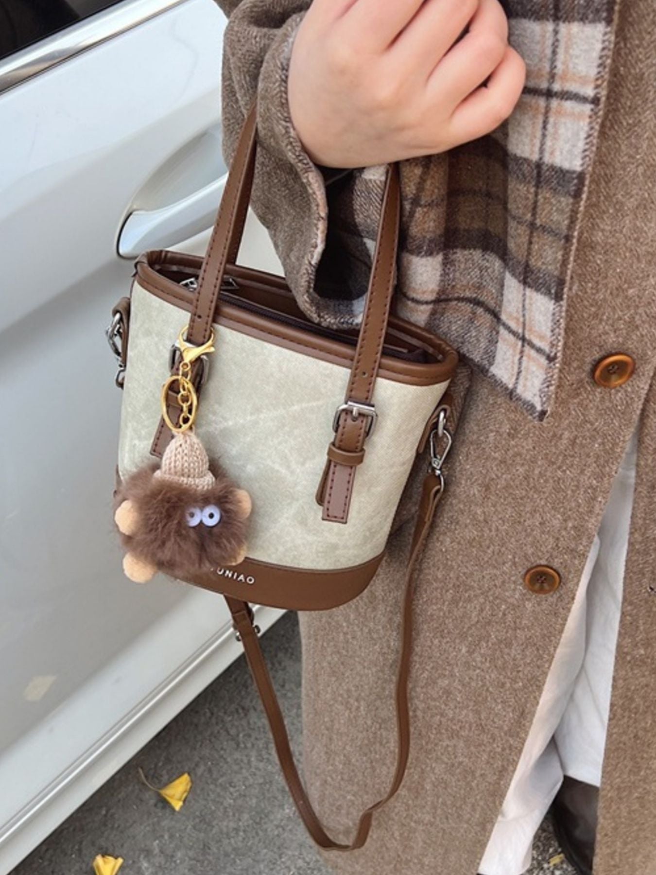 Person holding a beige handbag with a brown strap, wearing a plaid scarf and coat.