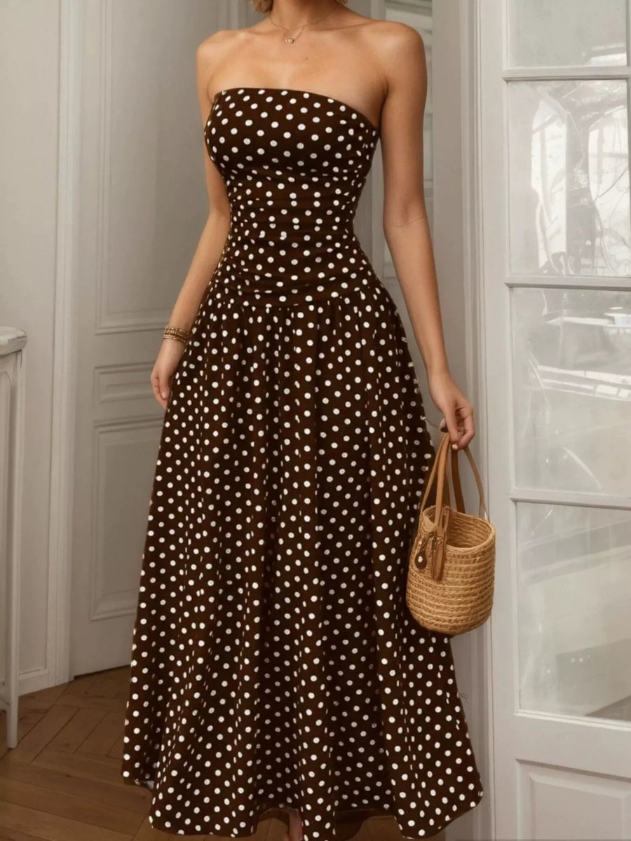 Woman wearing a brown strapless dress with white polka dots, holding a woven bag indoors.