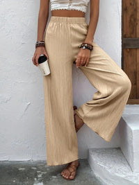 Person wearing beige wide-leg pants and a white top, holding a coffee cup against a white wall.