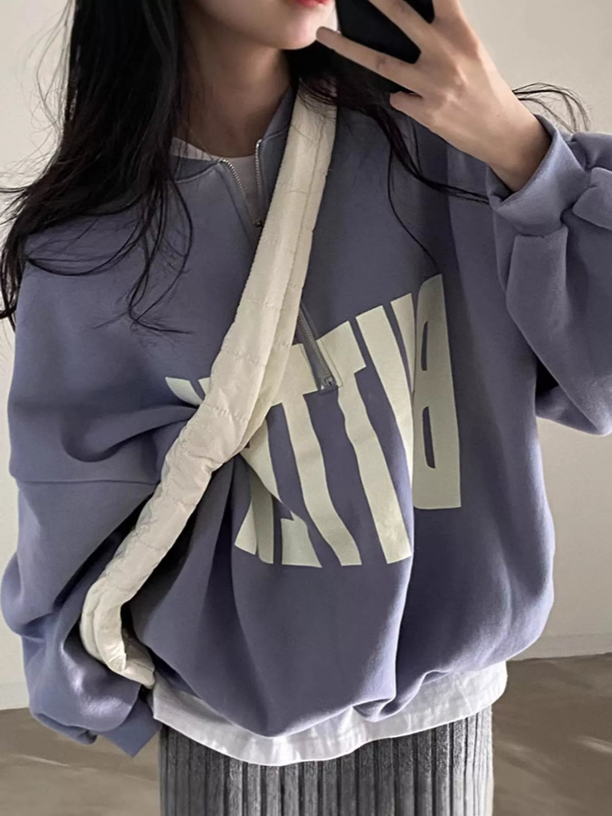 A person wearing an oversized lavender sweatshirt with a large cream graphic print on the front, layered over a white shirt and paired with a gray ribbed skirt. A cream quilted crossbody bag is also visible.