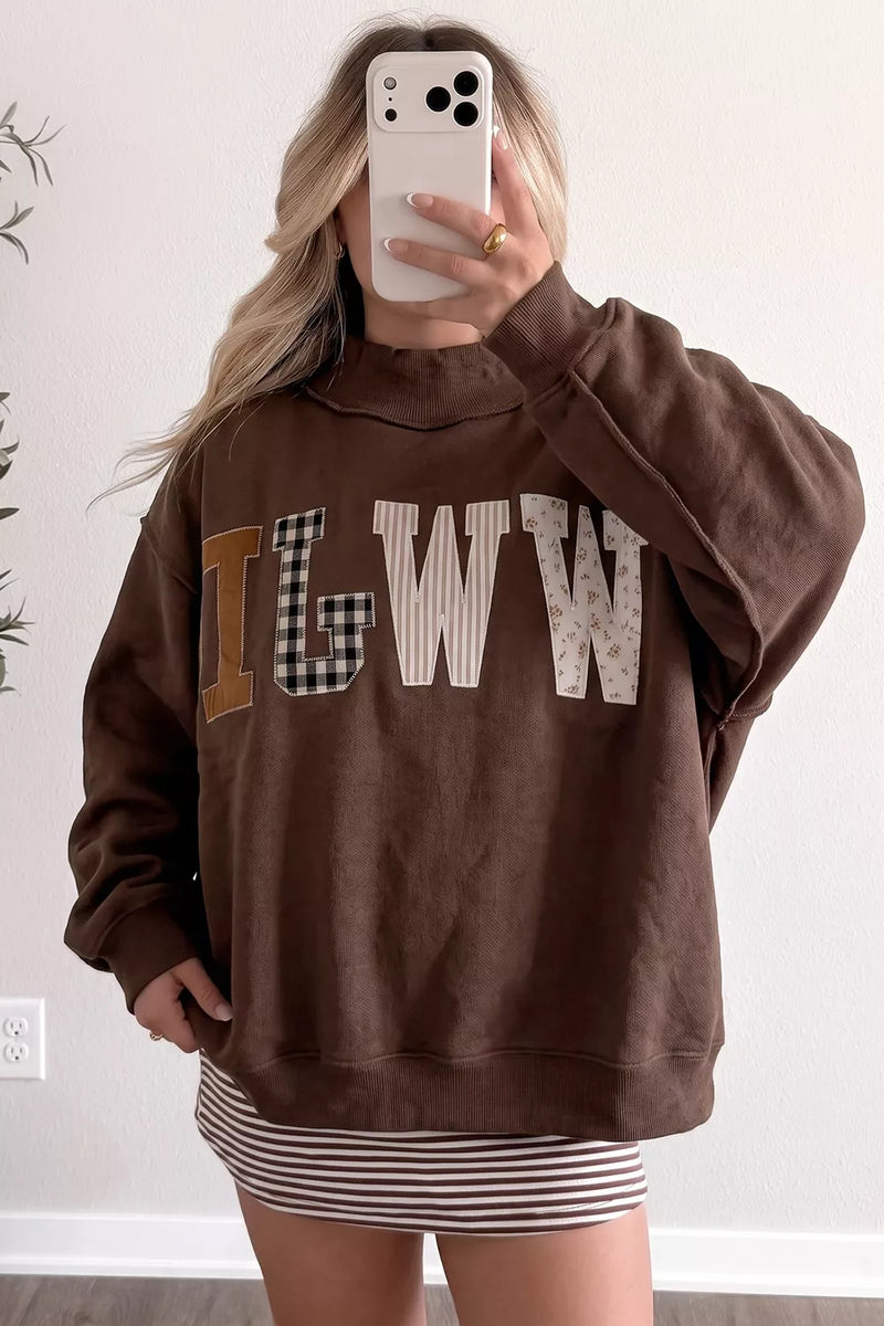 A woman takes a mirror selfie wearing a brown sweatshirt with large patchwork-style letters spelling 'ILGWW' across the chest, paired with a striped mini skirt.