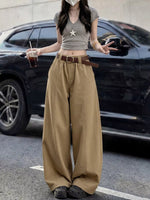 A person standing outdoors in front of a black SUV, wearing a gray cropped top with a star pendant, wide-leg tan pants, a brown belt, and dark shoes, while holding an iced drink.