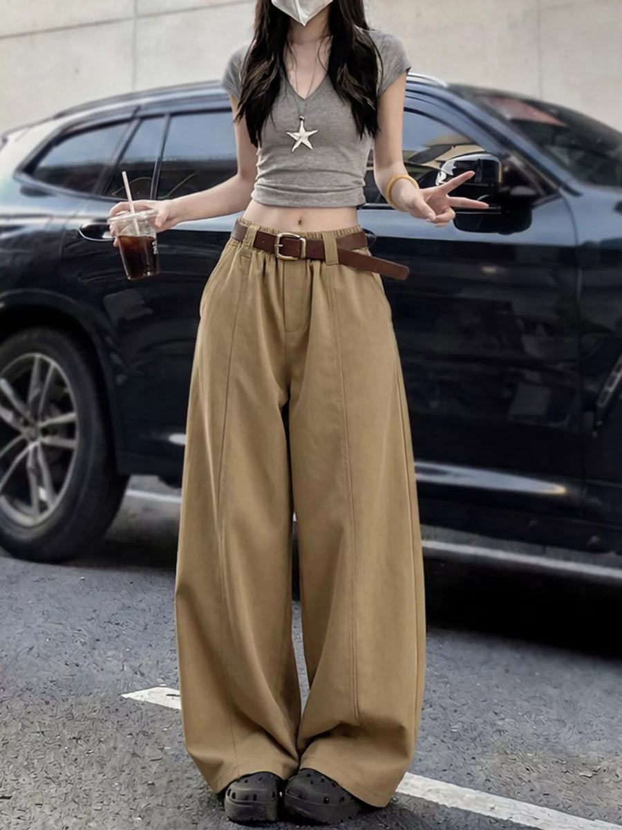 A person standing outdoors in front of a black SUV, wearing a gray cropped top with a star pendant, wide-leg tan pants, a brown belt, and dark shoes, while holding an iced drink.