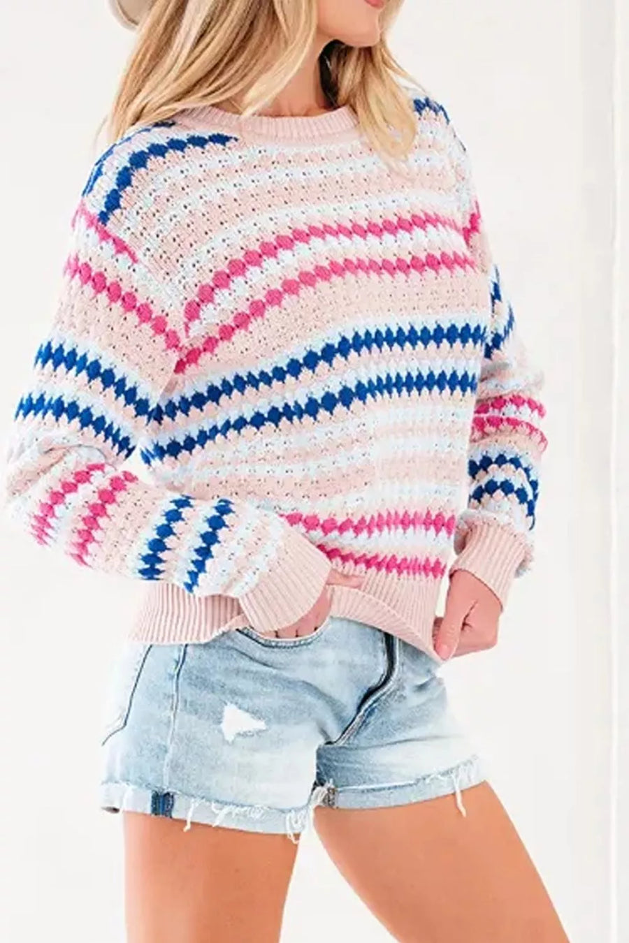 A woman wearing a chunky knit crewneck sweater with horizontal wavy stripes in pink, blue, and white, paired with light-wash denim shorts.