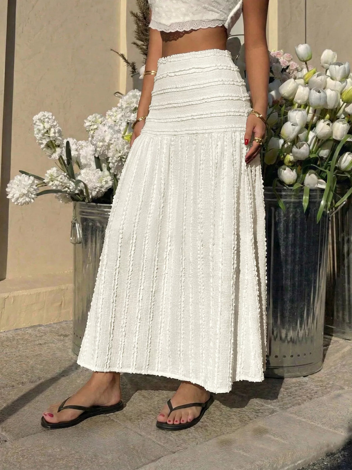 Person wearing a white outfit with a long skirt standing between two flower pots.