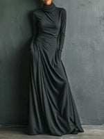 A woman wearing a long-sleeve, high-neck, floor-length black dress with gathered draping at the waist, standing against a dark textured wall.