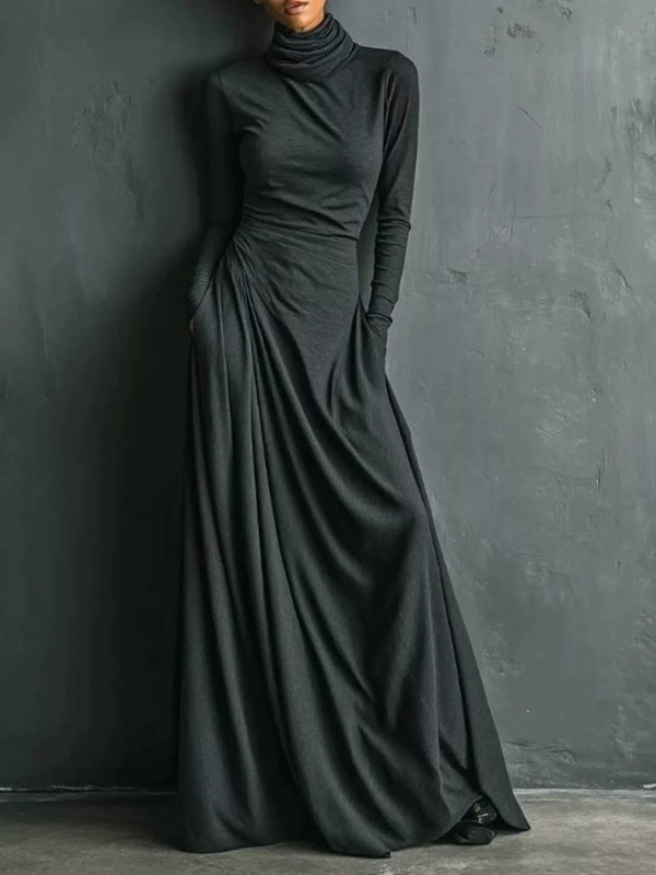 A woman wearing a long-sleeve, high-neck, floor-length black dress with gathered draping at the waist, standing against a dark textured wall.