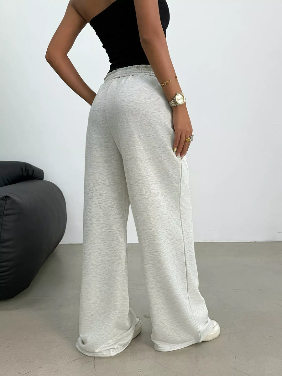 Person wearing gray wide-leg pants and a black top against a white wall.