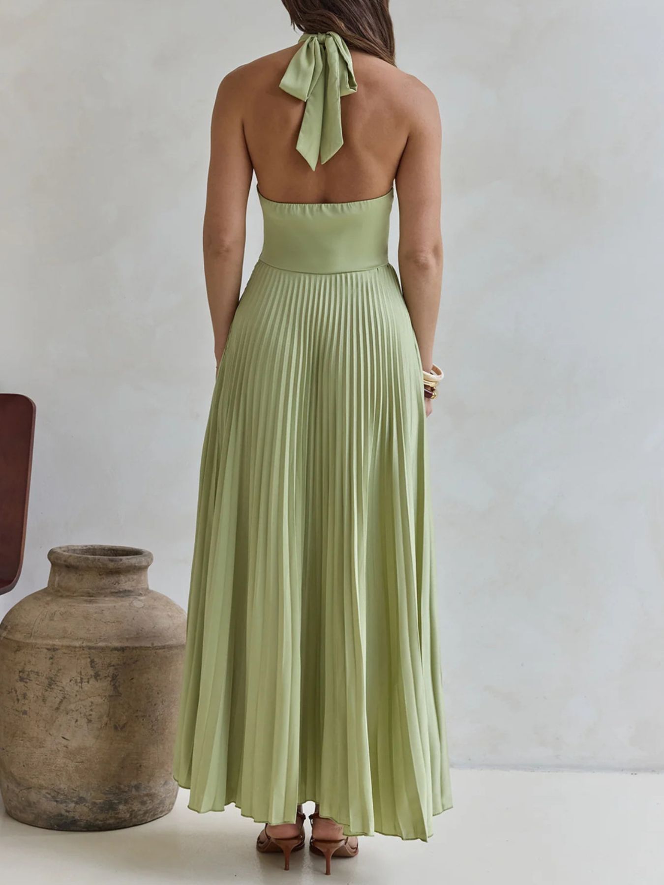 Woman wearing a long green dress with a halter neck and pleated skirt, standing against a plain background.