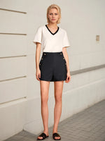 Woman wearing a white top and black shorts with button details against a light-colored wall.