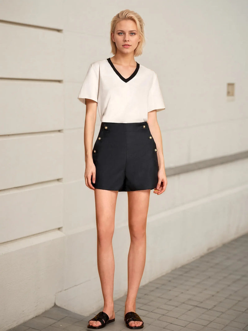 Woman wearing a white top and black shorts with button details against a light-colored wall.