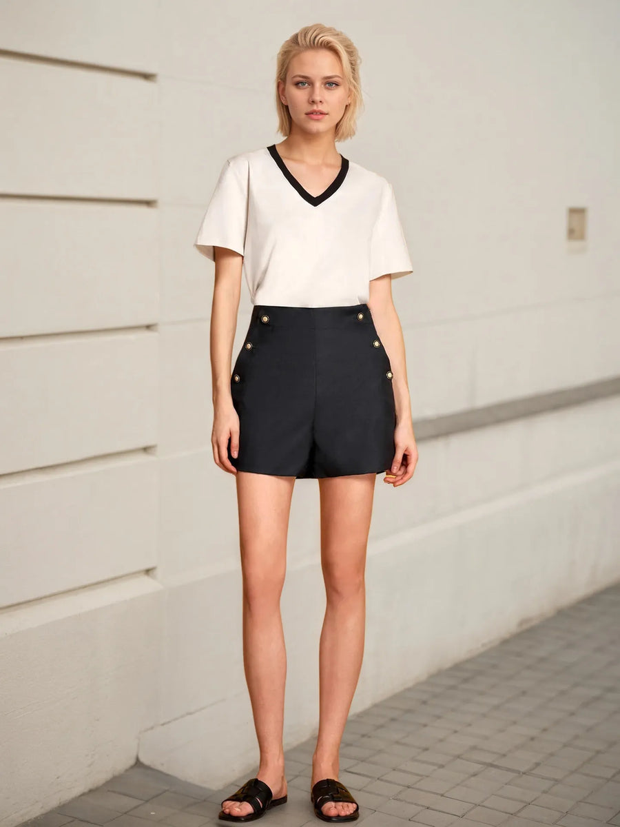 Woman wearing a white top and black shorts with button details against a light-colored wall.