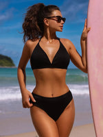 Woman in a black bikini standing on a beach with a surfboard.
