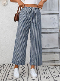 Person wearing blue and white striped pants with a white top and white sneakers, standing against a light wooden panel background.