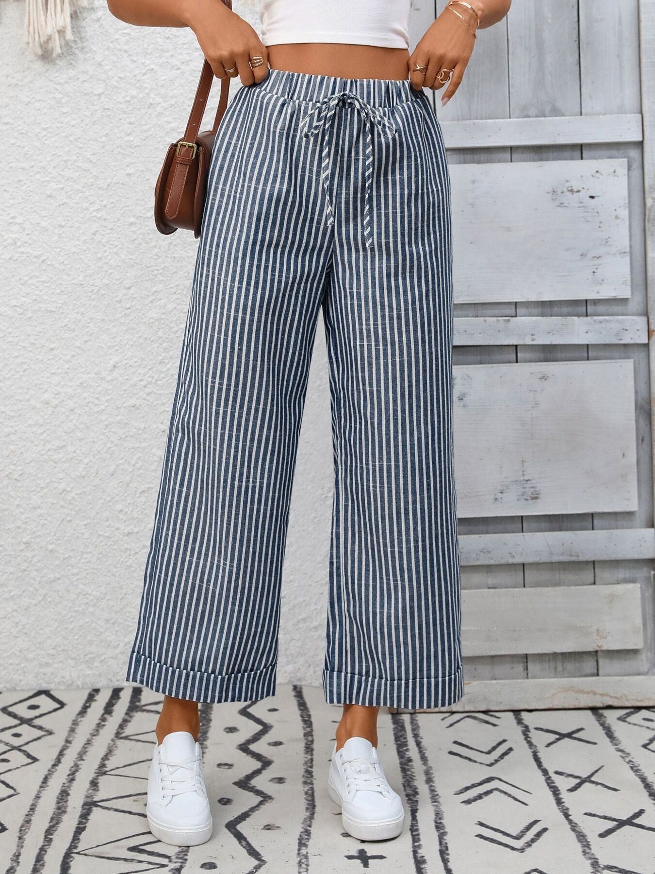 Person wearing blue and white striped pants with a white top and white sneakers, standing against a light wooden panel background.