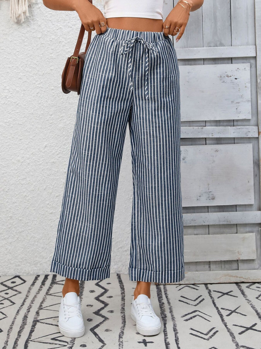 Person wearing blue and white striped pants with a white top and white sneakers, standing against a light wooden panel background.