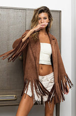 Woman wearing a brown suede jacket with fringe, white top, and denim shorts in front of a neutral background.