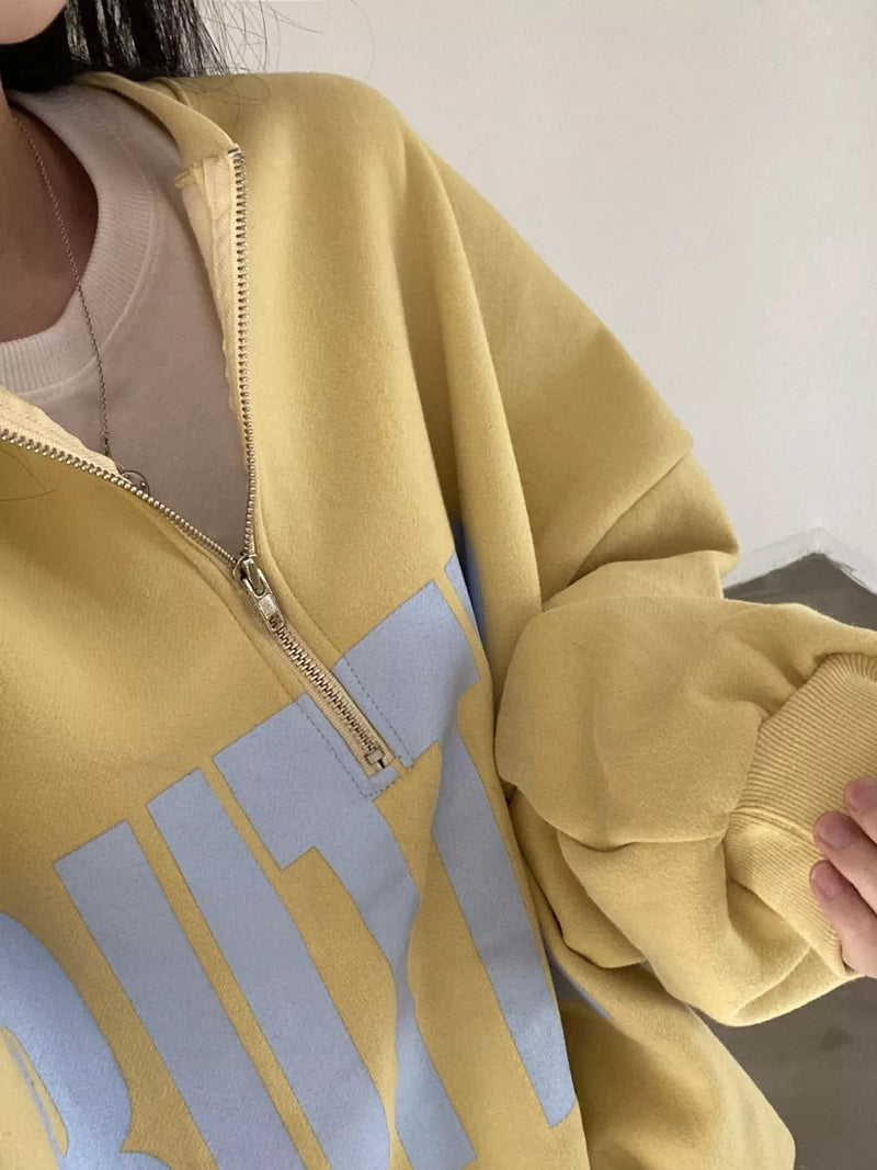 A close-up of a person wearing an oversized yellow quarter-zip sweatshirt with a large light blue graphic print on the front, layered over a beige t-shirt.