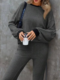 Person wearing a gray ribbed sweater and pants holding a white cup against a gray wall.