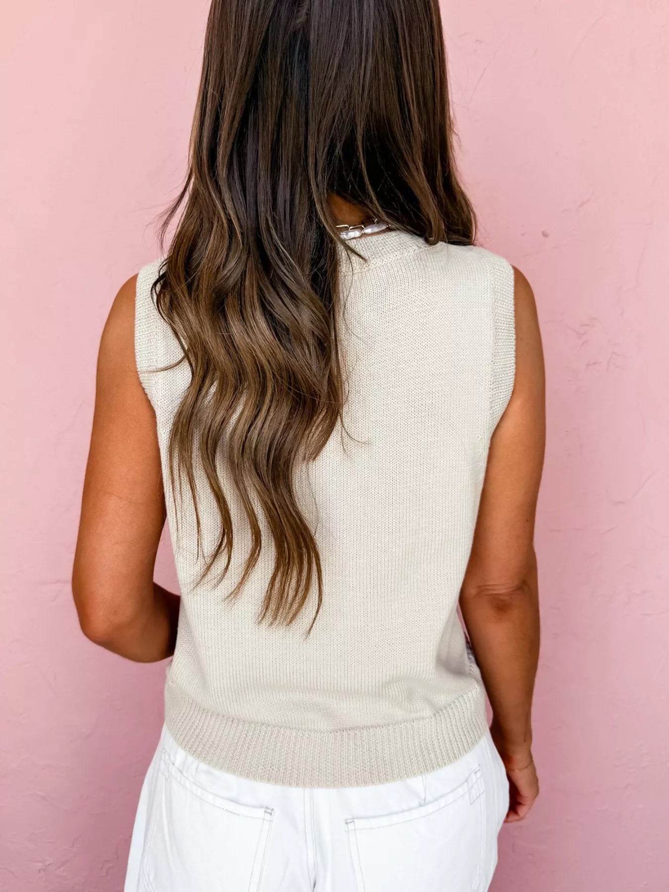 Person wearing a beige sleeveless sweater against a pink background